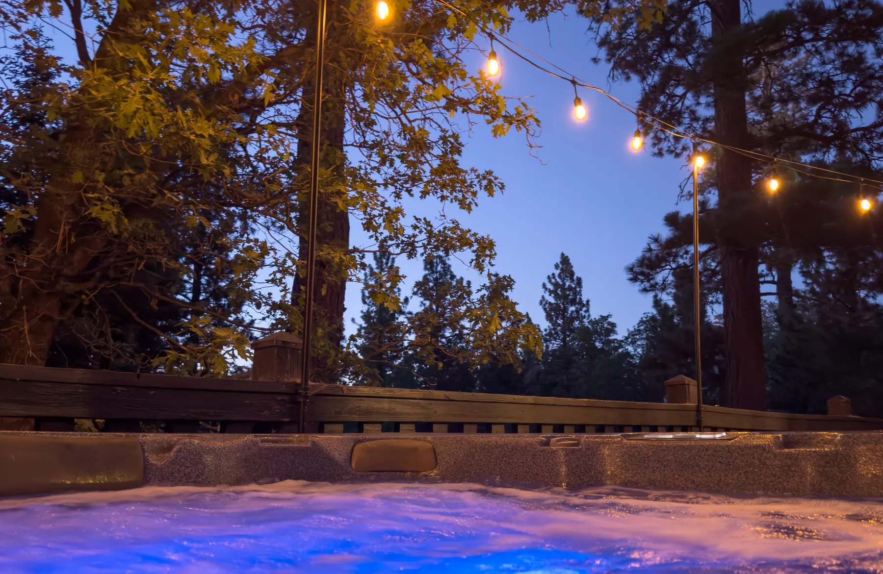 Private hot tub on the deck at Pine Grove Cabin at dusk, lit by string lights and surrounded by pine trees